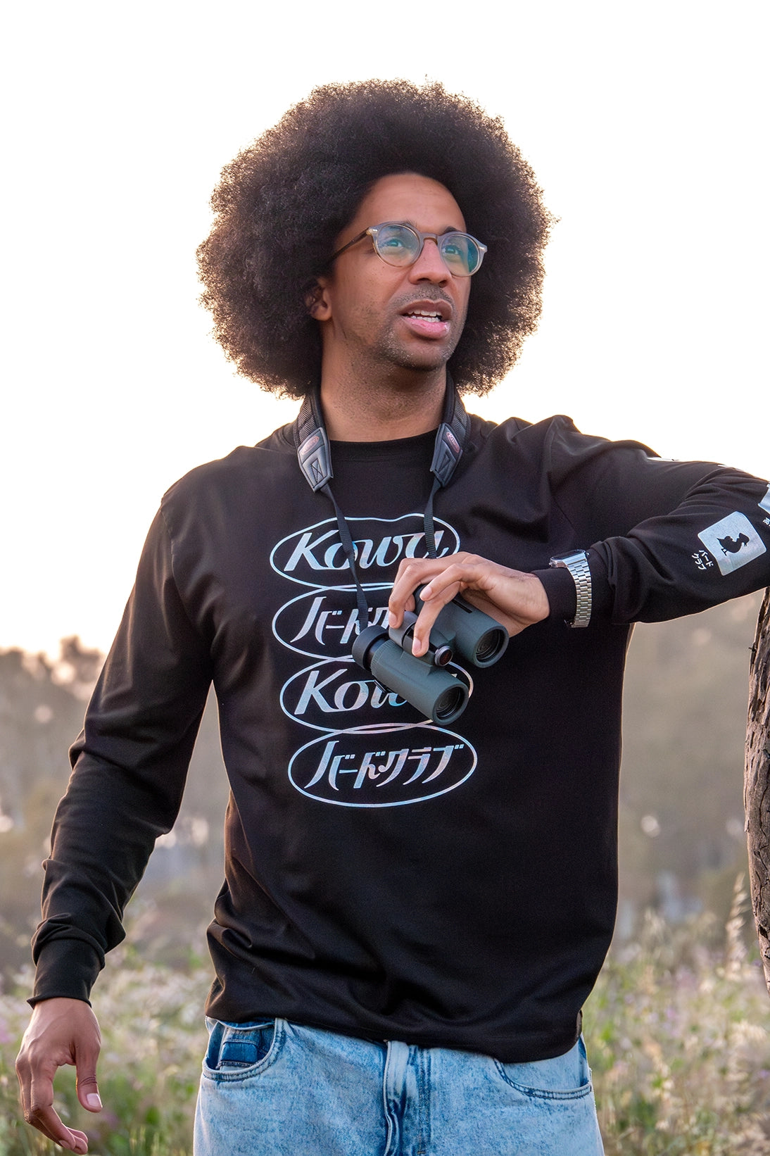 A man engages in bird watching, wearing a black long-sleeve T-shirt from the Bird Club and Kowa collaboration. The shirt features an iridescent gradient design with the Kowa logo and Japanese text "Bird Club" repeated vertically on the front. The left sleeve showcases icons related to bird watching, including fluorite crystal, waterproof design, high-resolution lens, and ornithology. The sustainable brand focuses on eco-friendly fashion and nature appreciation.