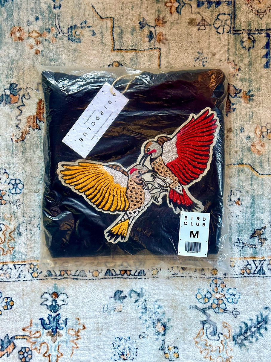 Bird Club sweatshirt featuring a vibrant embroidery of two Northern Flicker woodpeckers, packaged with sustainable materials. This image highlights Bird Club’s commitment to eco-friendly practices and ornithology-inspired apparel, making it a perfect choice for birding enthusiasts. Designed for both style and function, this sweatshirt stands out among bird watchers gifts and offers a thoughtful option for gifts for bird lovers who value nature, conservation, and high-quality clothing.