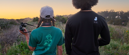 Two individuals stand in a field at sunset, both wearing shirts from the Bird Club and Kowa collaboration. The woman on the left wears a green T-shirt featuring a mint green line drawing of a bird and the text "Bird Club" on the back. She holds binoculars and has a Kowa strap around her neck. The man on the right wears a black long-sleeve shirt with the same bird illustration and "Bird Club" text on the back.