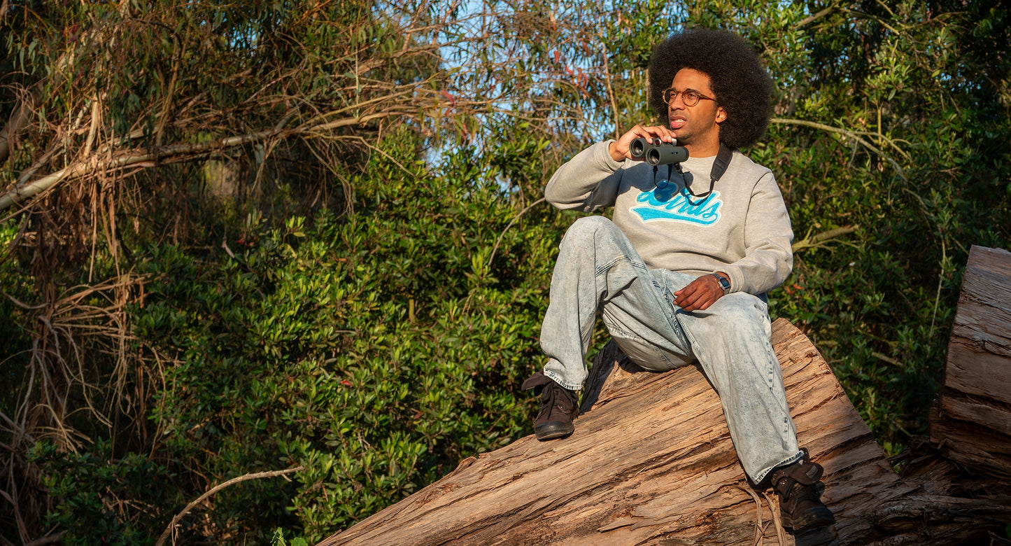  A man sits atop a large fallen tree trunk in a dense forest, holding binoculars as he engages in bird watching. He is wearing a gray Bird Club sweatshirt featuring the word "Birds" in eye-catching blue chenille embroidery. The sweatshirt is part of a sustainable brand collection, emphasizing the intersection of eco-friendly fashion and ornithology. The natural setting, with lush green foliage surrounding him, highlights the brand's connection to nature and the outdoors.