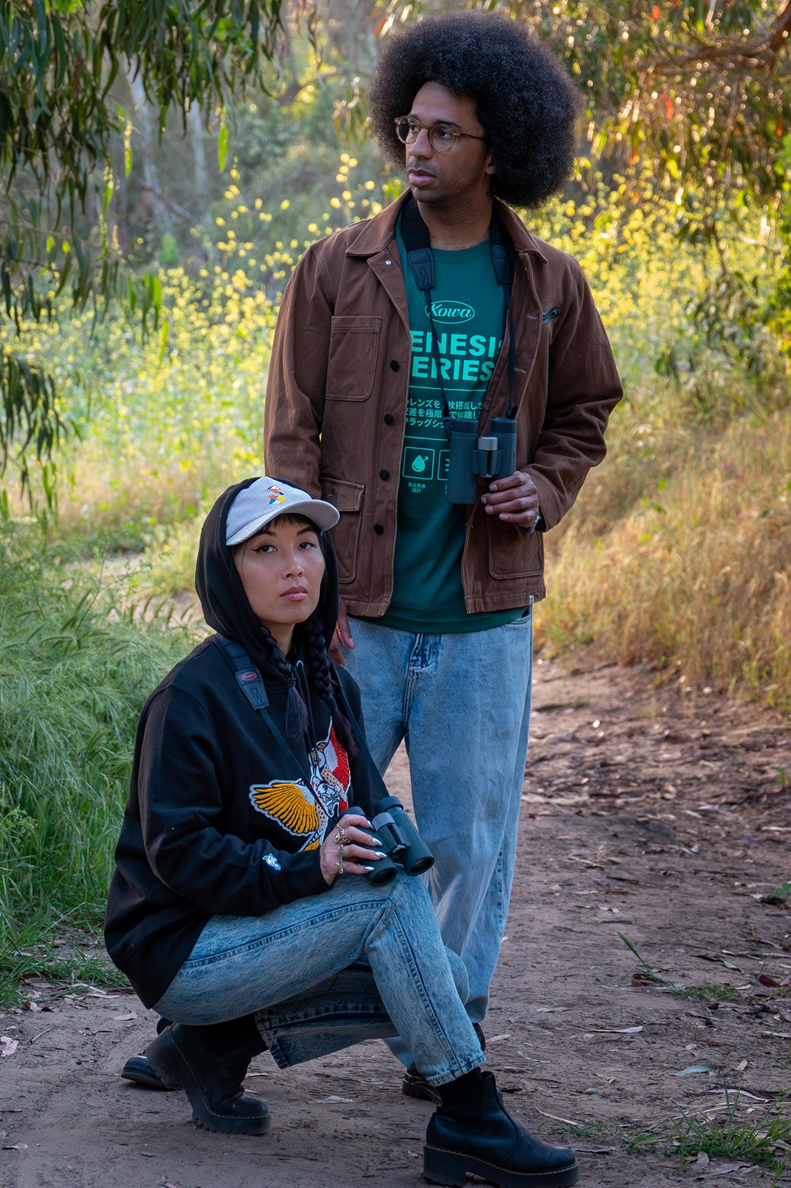 Two birding enthusiasts, a woman and a man, engage in bird watching in a scenic, natural environment. The woman, kneeling with Kowa binoculars, wears a BIRD CLUB hoodie, while the man stands with binoculars around his neck and a Kowa Genesis Series shirt. This image emphasizes ornithology, birding, and sustainable fashion, showcasing the naturalist lifestyle promoted by BIRD CLUB's commitment to high-quality, eco-friendly products