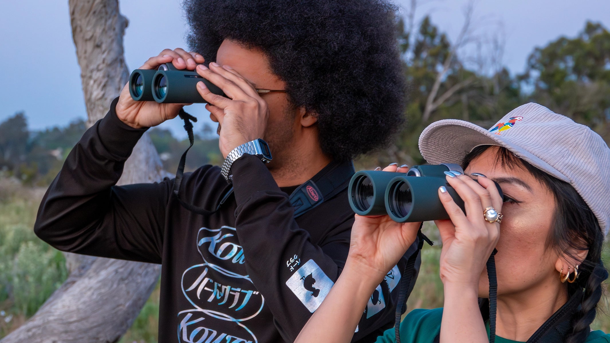 Two naturalists using Kowa binoculars for bird watching. The man wears a black BIRD CLUB shirt and the woman sports a cap with the BIRD CLUB Mandarin Duck Logo, both deeply engaged in ornithology. This image highlights the birding activity promoted by the BIRD CLUB, emphasizing the sustainable brand's dedication to high-quality, eco-friendly products.