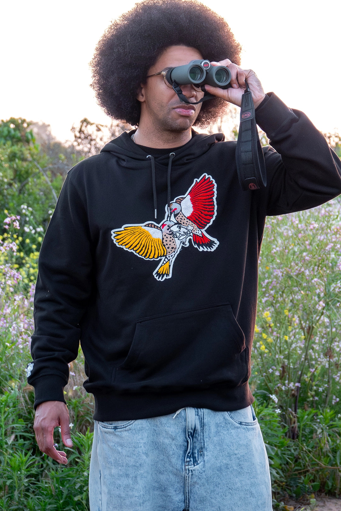 Dexter Thomas, wearing a BIRD CLUB hoodie with embroidered Northern Flicker woodpeckers, uses binoculars for bird watching in a natural setting. This image highlights the ornithology and naturalist interests promoted by BIRD CLUB, emphasizing the brand's commitment to sustainable practices and eco-friendly apparel.