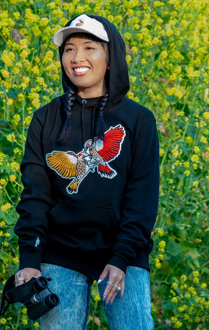 A smiling Mamiko wears a BIRD CLUB hoodie featuring embroidered Northern Flicker woodpeckers and a cap with the BIRD CLUB logo, holding binoculars. This image highlights the brand's commitment to sustainable practices and appeals to bird watching and ornithology enthusiasts, reflecting BIRD CLUB's dedication to eco-friendly and high-quality apparel.