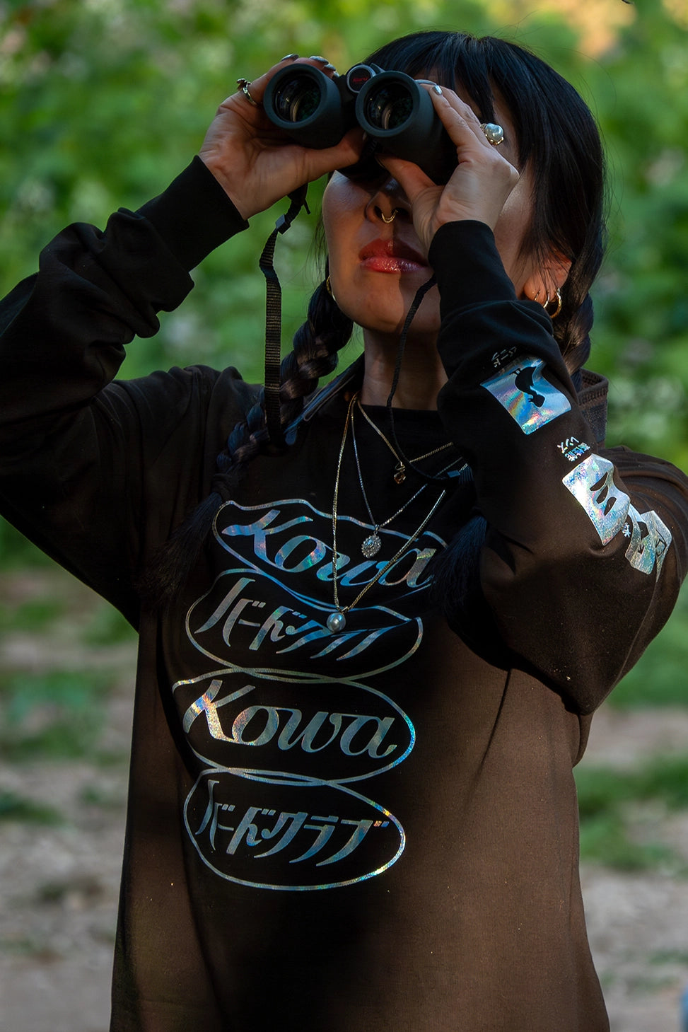 Mamiko, wearing a BIRD CLUB and Kowa long-sleeve t-shirt, uses binoculars for bird watching in a natural setting. This image highlights her involvement in ornithology and naturalist activities, reflecting the values promoted by BIRD CLUB. It emphasizes the brand's commitment to sustainable practices and eco-friendly apparel