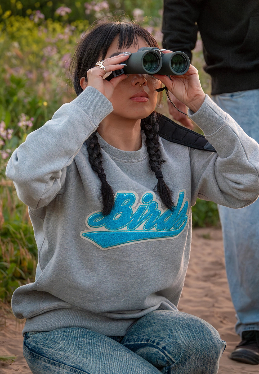 A woman kneels in an outdoor setting, using binoculars to bird watch. She wears a gray Bird Club sweatshirt featuring a stylish chenille embroidery of the word "Bird" in a vibrant blue. The sweatshirt, part of a sustainable brand collection, perfectly embodies the passion for ornithology. The image highlights the connection between sustainable fashion and bird watching, emphasizing the brand's commitment to eco-friendly apparel for bird enthusiasts.