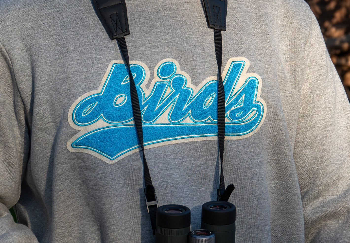 Close-up of a Bird Club sweatshirt featuring chenille embroidery of the word 'Birds' in blue, framed to highlight the detailed texture and quality of the design. Binoculars hang around the wearer's neck, emphasizing Bird Club's dedication to sustainable brands, bird watching, and ornithology.