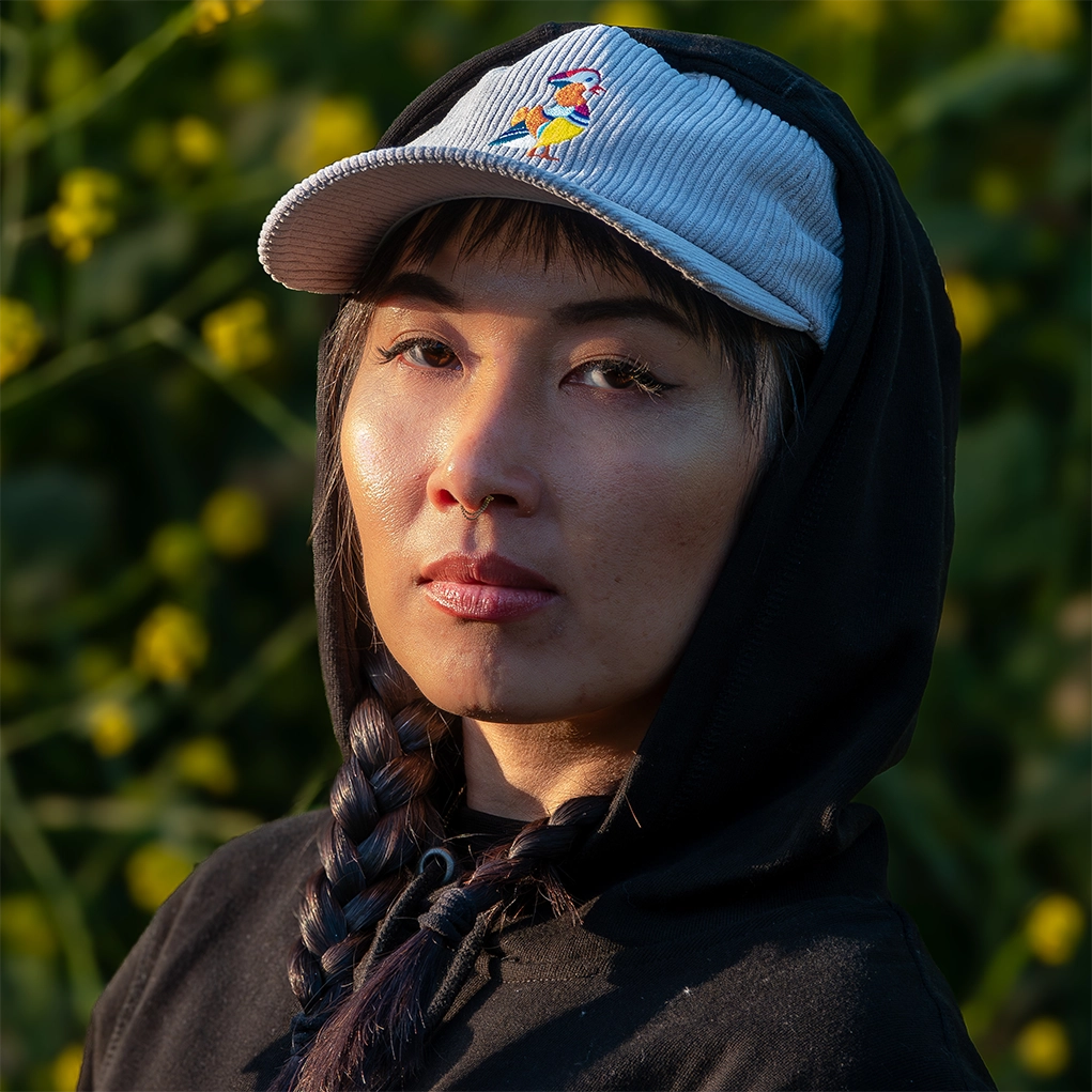 A woman wearing a gray cap with an embroidered bird logo, representing Bird Club, and a black hoodie, a part of the bird club clothing collection. The ornithology shirt emphasizes her love for birding. The background features lush greenery, adding a natural and serene touch to the scene.