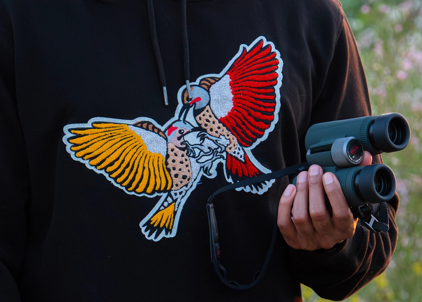 A close-up of a person holding binoculars while wearing a black Bird Club sweatshirt adorned with a vivid chenille embroidery of two woodpeckers, one with yellow and the other with red wings, locked in a dynamic pose. The detailed embroidery emphasizes the sustainable brand's commitment to ornithology and bird watching. This eco-friendly apparel piece is designed for bird enthusiasts and nature lovers, blending stylish design with environmental consciousness.