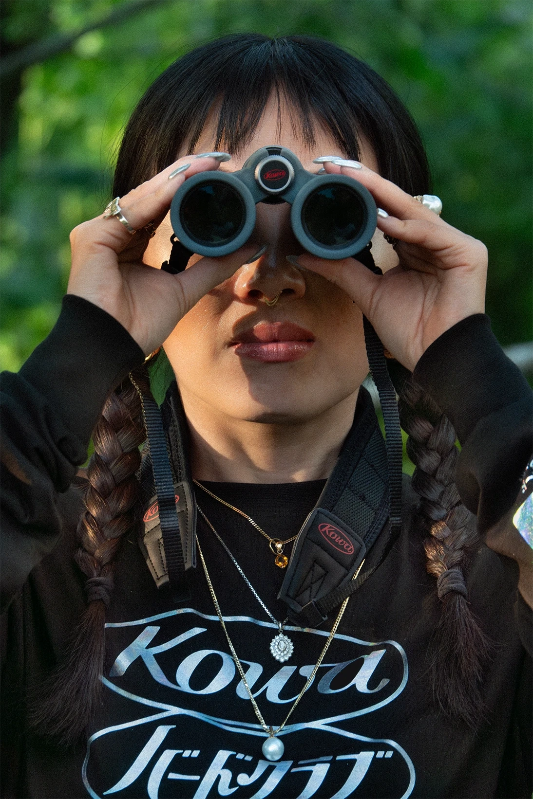 A woman wearing a Kowa and BIRD CLUB-branded shirt and necklace uses Kowa binoculars for bird watching in a lush, green environment. The image highlights her connection to ornithology, birding, and her role as a naturalist. Kowa optics and apparel are part of a sustainable brand dedicated to high-quality, ethical products