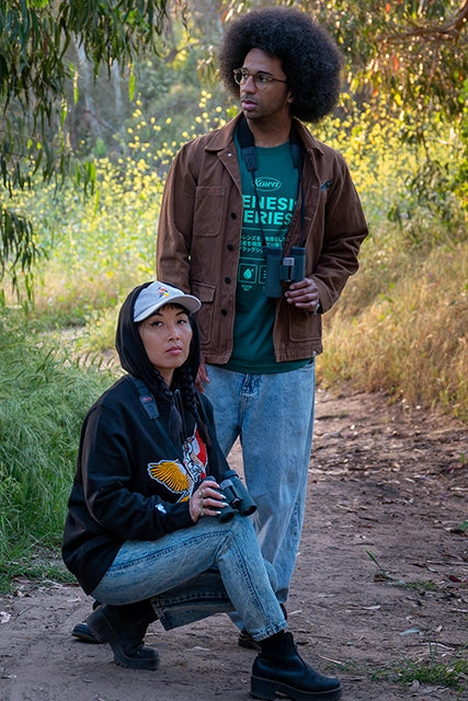 A man and woman engaged in bird watching in a forest, with the woman kneeling and the man standing. The woman wears a black Bird Club sweatshirt featuring a vibrant chenille embroidery of two Northern Flicker woodpeckerswith yellow and red wings. The man wears a brown jacket over a green T-shirt with binoculars around his neck. Both are part of a sustainable brand collection, highlighting the connection between eco-friendly fashion and ornithology. 