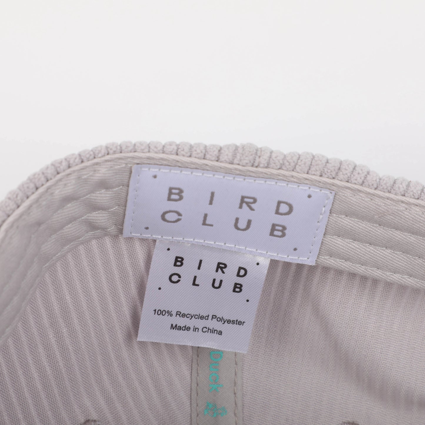  A close-up view of the inside of a recycled corduroy hat from Bird Club, showing the label and tags. The tags display the Bird Club logo and information about the hat being made from 100% recycled polyester. The light gray corduroy fabric adds a textured and eco-friendly element to the hat. This sustainable brand emphasizes bird watching and ornithology, appealing to environmentally conscious consumers. 