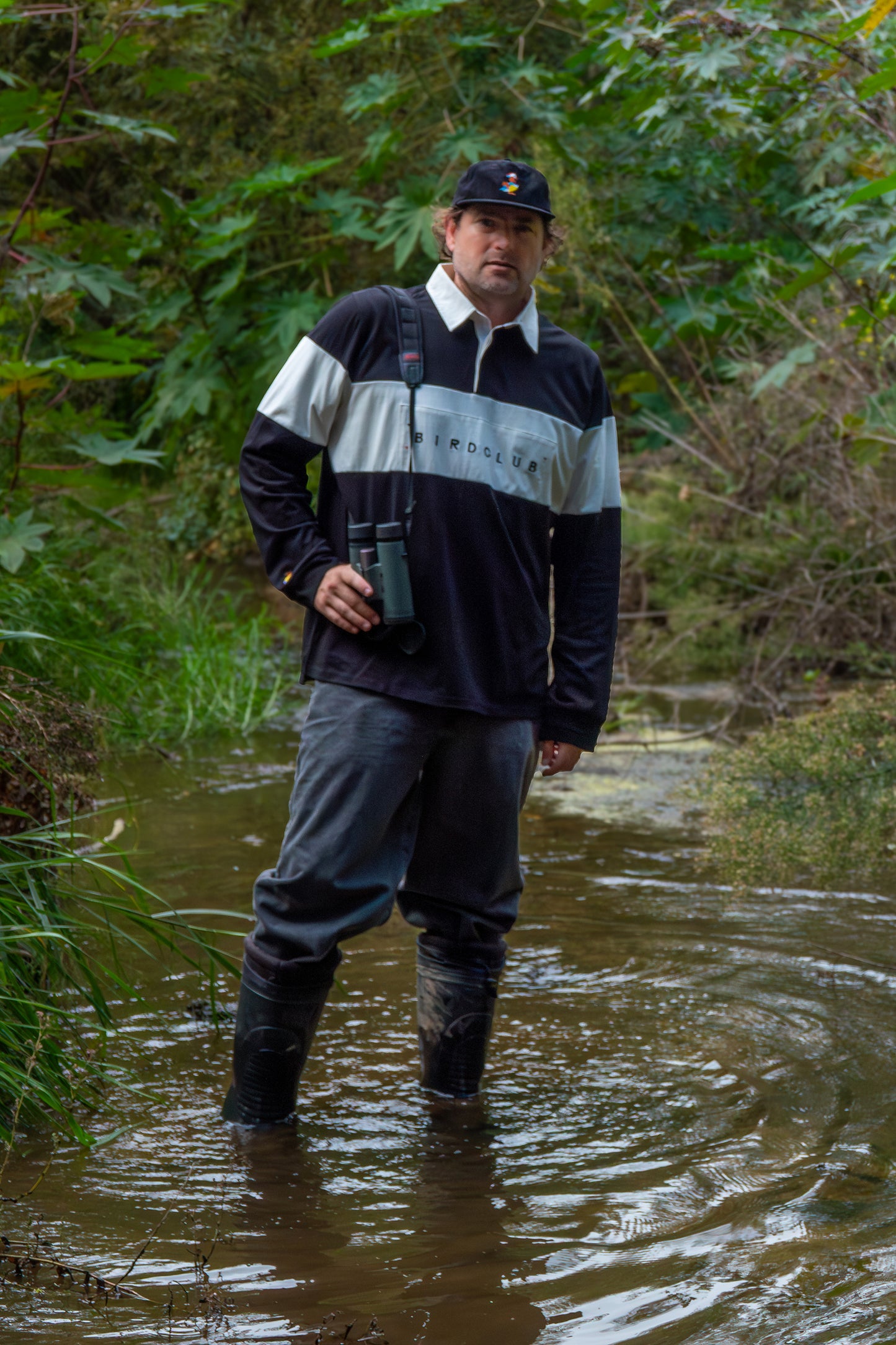 Man in nature wearing Bird Club apparel, standing in a creek with binoculars, embodying the perfect look for gifts for bird lovers and gifts for birders. Representing sustainable brands, Bird Club combines stylish, eco-friendly fashion with a passion for birdwatching, ideal for nature enthusiasts.